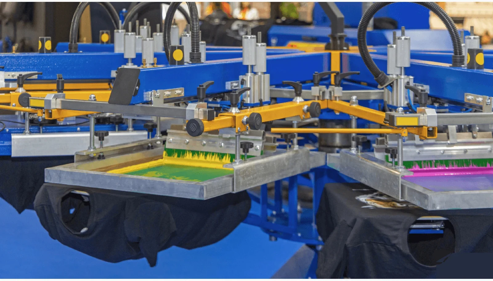 Custom merchandise production UK using an automatic screen-printing press with yellow and magenta screens over black T-shirts.