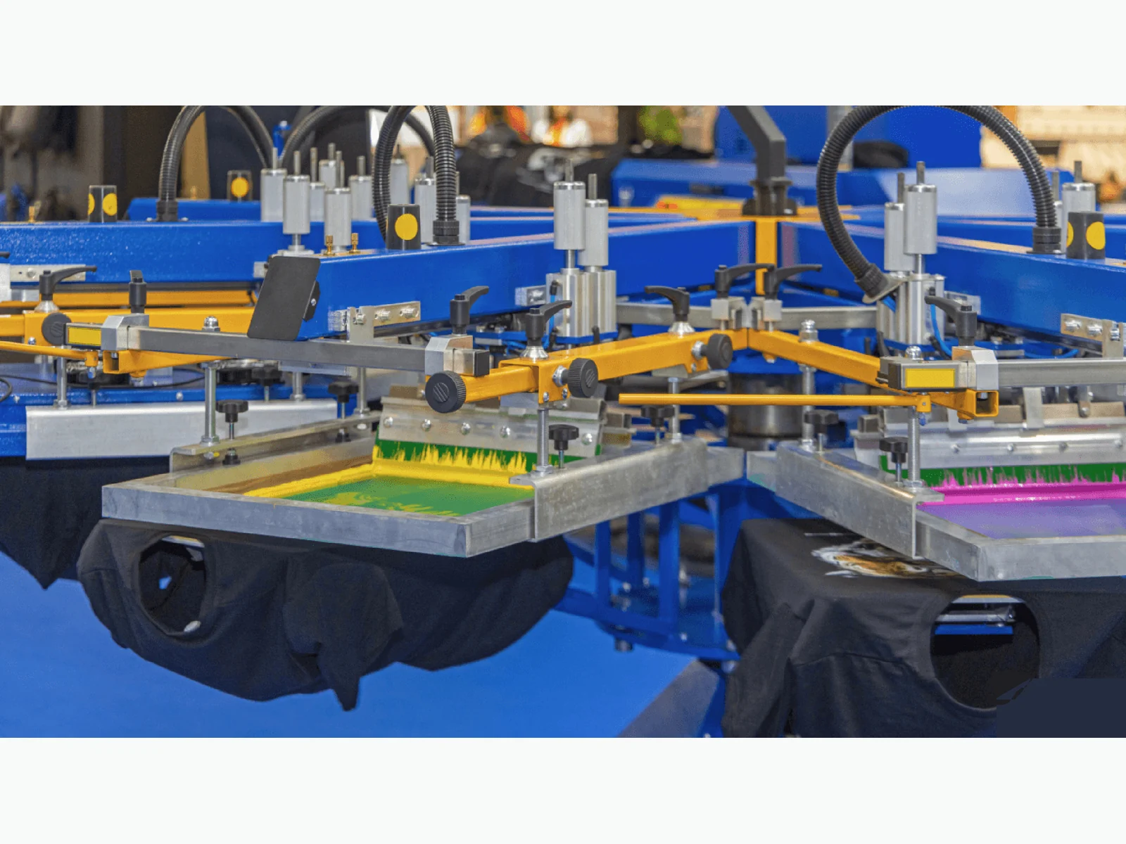 Automatic screen-printing press with yellow and magenta screens over black T-shirts.